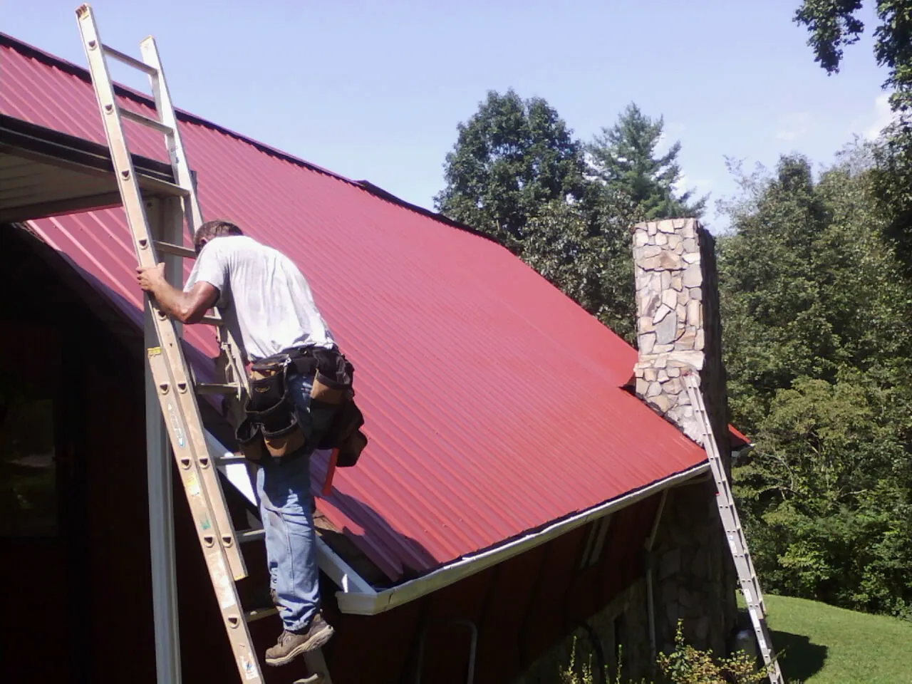 Roofer on a ladder beside a red metal roof and stone chimney — Soffit & Fascia Repair by Roofing Brothers in Flowery Branch